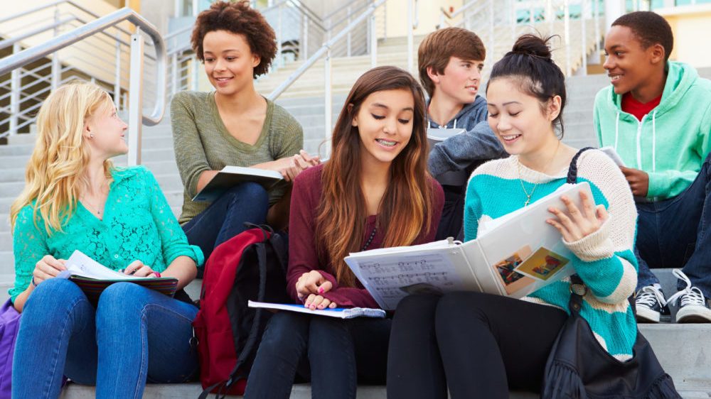 Group of high school students sitting outside building of one of the best schools their parents could find when moving.