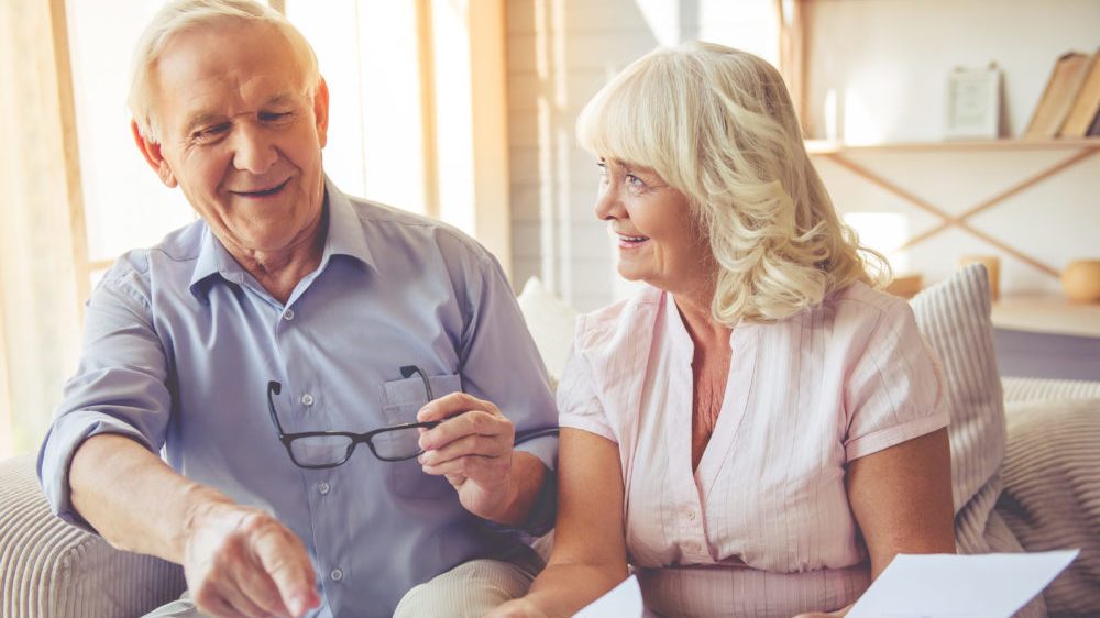 Handsome old man and beautiful woman are discussing home resizing project and smiling while sitting on couch.