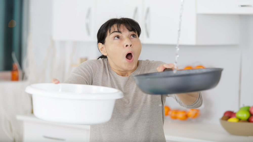 Surprised woman holding bucket while water droplets leak from ceiling. She is wondering whether her homeowner's policy will cover the damage.