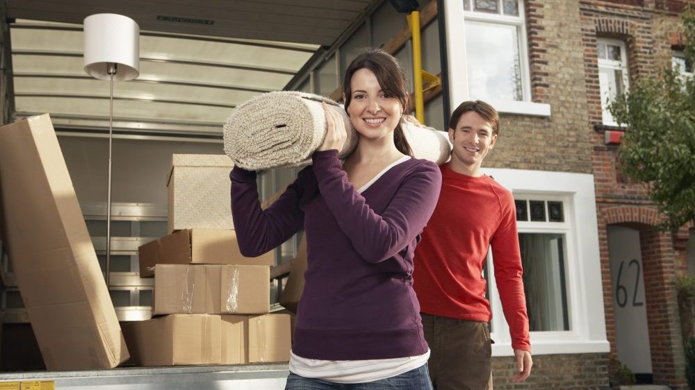 Portrait of happy young couple carrying rolled carpet from a rental truck that's insured.