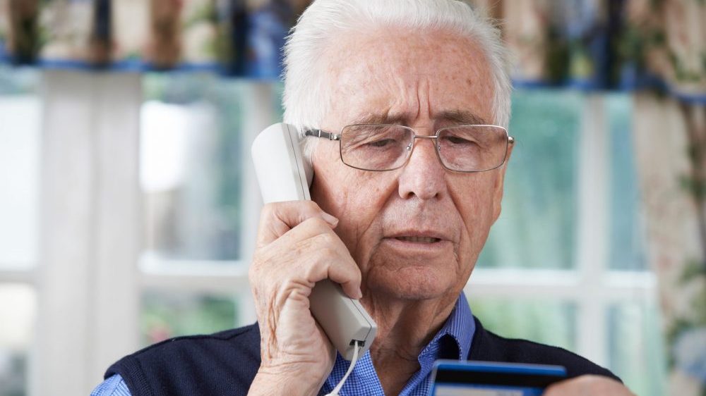 Senior man giving credit card details on a landline phone to a fraudulent robocall.