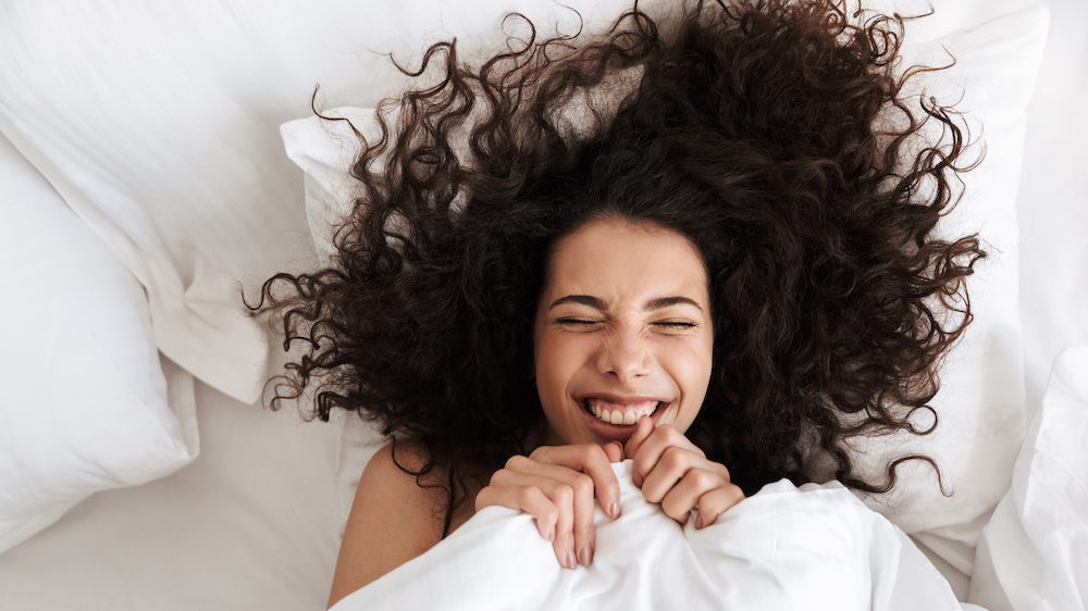 Portrait from above of cheerful woman with dark curly hair and a smiling face lying in bed under white bed sheets pulled up to her chin.