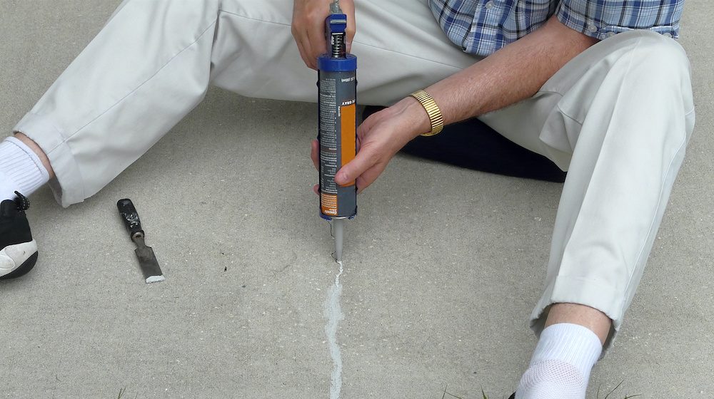 Man filling a cracked concrete in patio block with a caulking gun and putty knife.