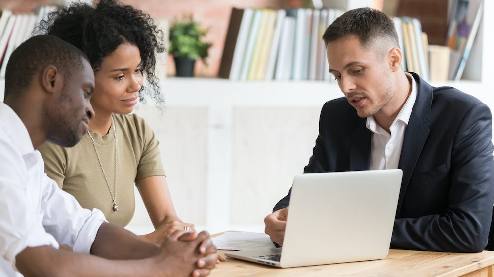 Male real estate agent meeting with millennial couple showing properties on laptop. The agent can only answer questions allowed within Fair Housing Laws.