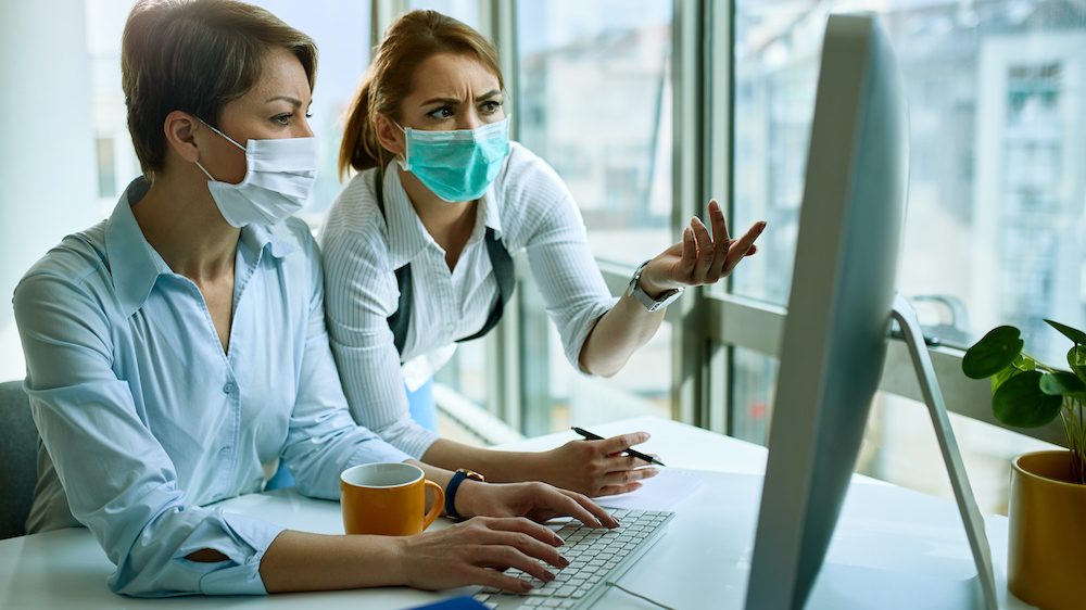 Female colleagues wearing face masks while working on a computer and communicating about possible coronavirus cyber scams via an e-mail.