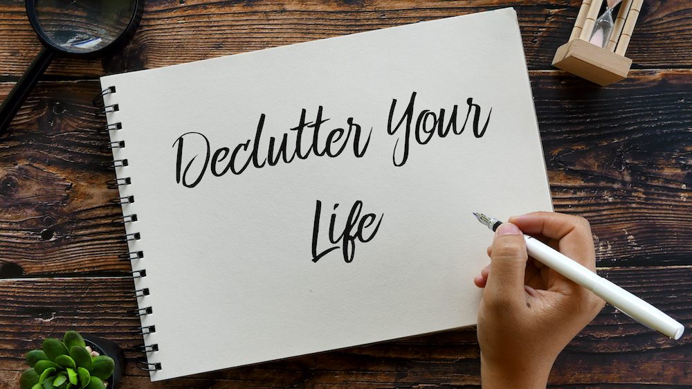 Top view of magnifying glass,sand clock, plant, pen with handwriting "Declutter Your Life" on notebook on wooden background. Theme of how to conquer clutter in your life.