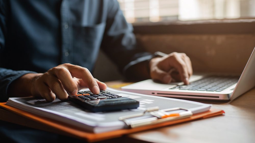 Gentleman's hand calculating the closing costs before selling his home at desk in an office.
