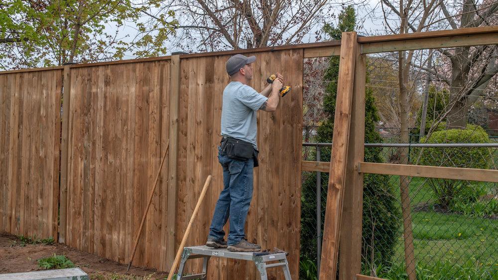 A white, middle-aged man builds a wooden fence in his backyard.