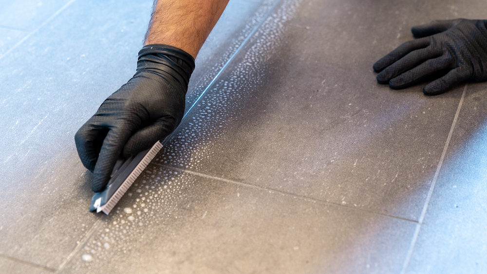 A close-up of a professional cleaner cleaning dirty tile grout with a brush blade and foamy soap on a gray bathroom floor.