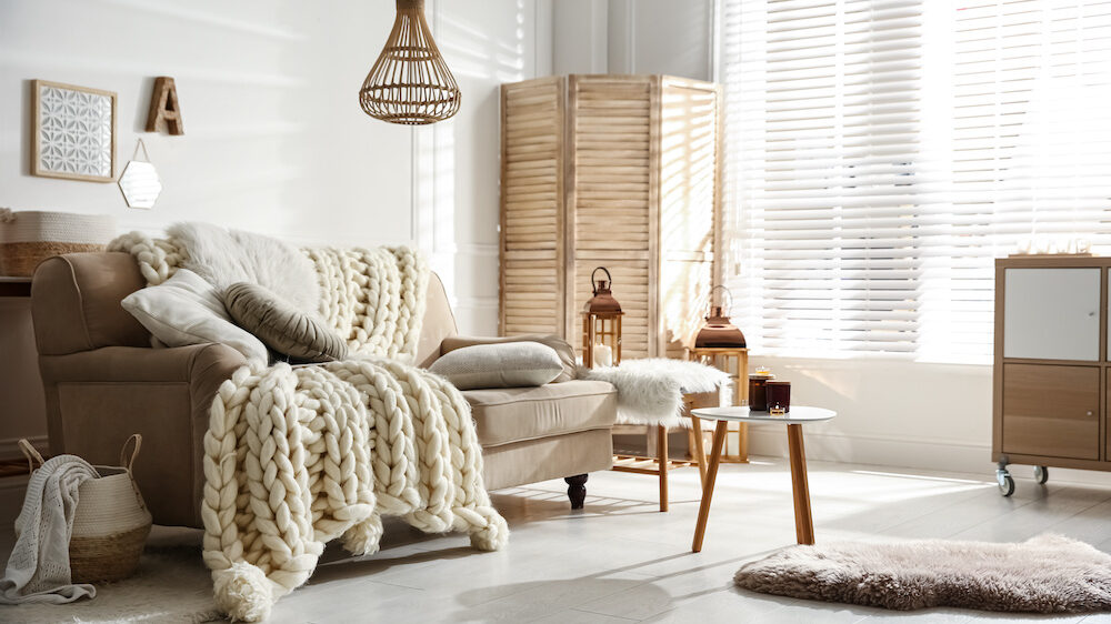 Cozy living room interior with beige sofa, knitted blankets, cushions, and off-white window blinds and shades.
